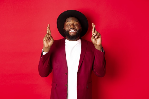 Hopeful african american man making wish, holding fingers crossed for good luck and smiling optimistic, standing against red party background