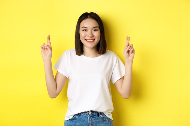 Hopeful asian girl making wish, cross fingers for good luck and praying with eyes closed, standing over yellow background.