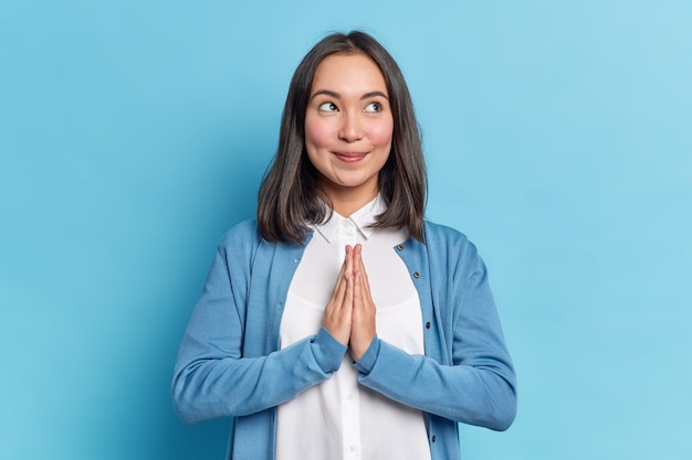 Hopeful beautiful Asian woman presses palms together prays for something makes supplicating namaste pray gesture dressed in neat clothes