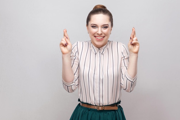 Hopeful beautiful young woman in striped shirt and makeup and ban hairstyle, standing with crossed fingers and looking at camera with toothy smile. indoor studio shot, isolated on grey background.