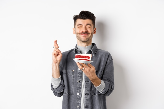 Hopeful birthday guy making wish with fingers crossed, holding bday cake on party, standing against white background.