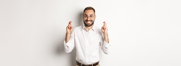 Hopeful businessman smiling and cross fingers for good luck making a wish standing over white backgr