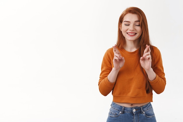 Hopeful cheerful redhead woman urban woman red haircut close eyes smiling joyfully cross fingers good luck faithfully awaiting wish come true anticipating white wall