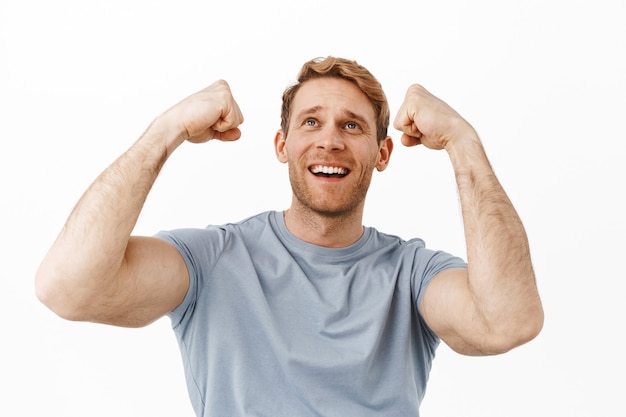 Hopeful handsome sports fan with red hair and strong fit body, raising hands up, achieve success, rejoicing and celebrating sport team score goal, standing over white wall