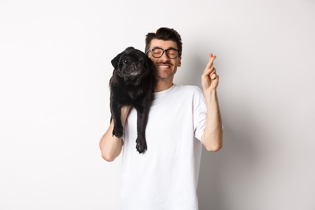 Hopeful smiling dog owner making a wish, holding cute black pug on shoulder and cross fingers for good luck, white background