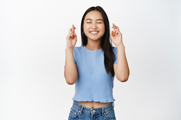 Hopeful smiling girl close eyes wishing something with crossed fingers praying and anticipating standing over white background