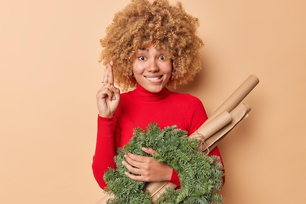 Hopeful woman bites lips keeps fingers crossed makes wish believes in good luck carries green spruce wreath and rolled paper isolated over beige background. New Year preparations and winter time