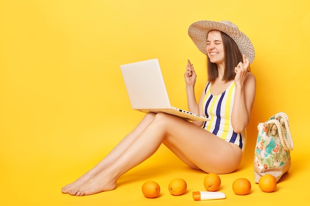Hopeful woman wearing striped onepiece swimsuit and straw hat posing isolated over yellow background booking tickets for vacation online praying to have free dares for departure