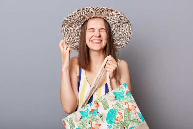 Hopeful woman wearing striped onepiece swimsuit and straw wide brim hat posing isolated on gray background holding bag closed eyes crossed fingers for good luck dreaming about resort