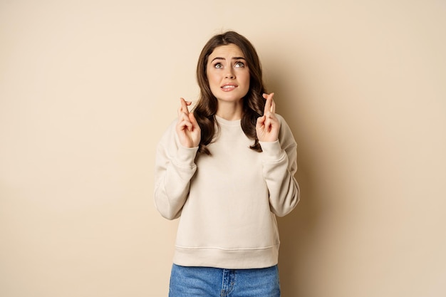 Hopeful worried girl cross fingers making wish standing over beige background