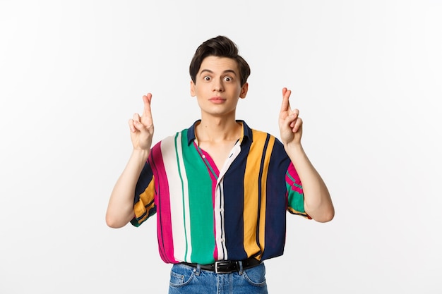 Hopeful young androgynous man cross fingers for good luck, making wish, waiting for something, standing over white.