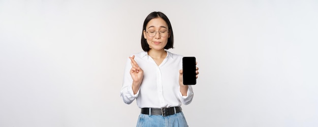 Hopeful young asian woman showing mobile phone screen app interface and fingers crossed hoping for smth making wish or paying standing over white background