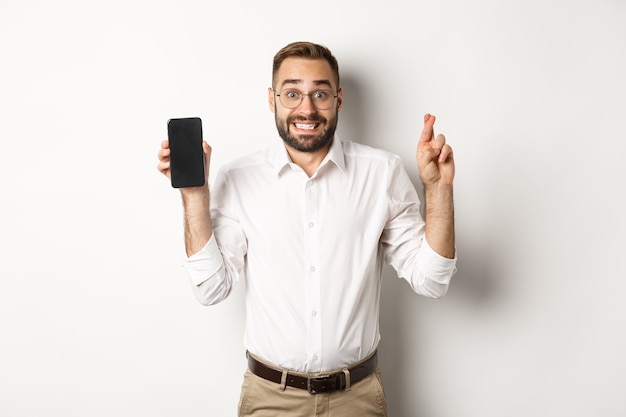 Hopeful young business man showing mobile screen, holding fingers crossed, waiting for online results, standing over white background