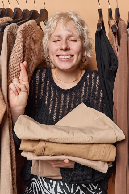 Фото Обнадеживающая молодая светловолосая женщина держит пальцы скрещенными верит в удачу держит глаза закрытыми держит сложенную одежду позирует в шкафу возлагает большие надежды на что-то широко улыбается концепция одежды