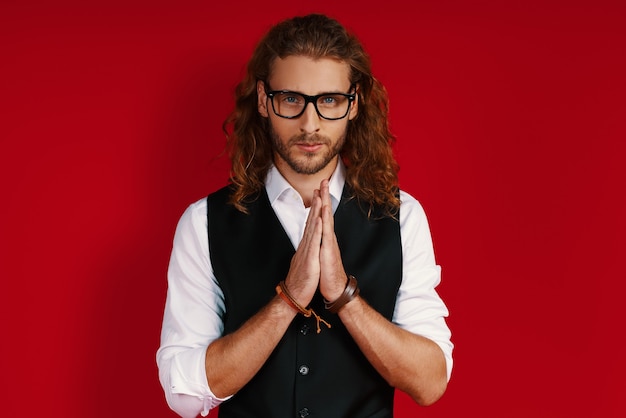 Hopeful young man in elegant clothing looking at camera and keeping hands clasped while standing against red wall