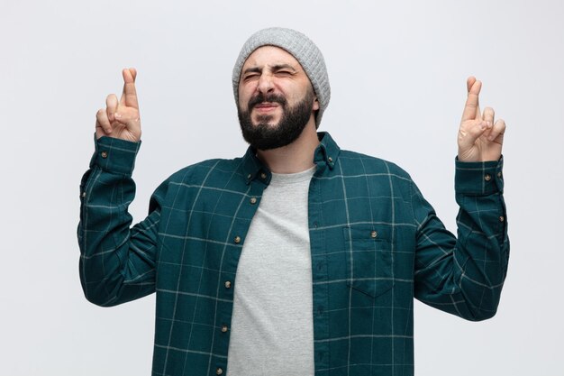 Hopeful young man wearing winter hat crossing fingers making wish with closed eyes isolated on white background