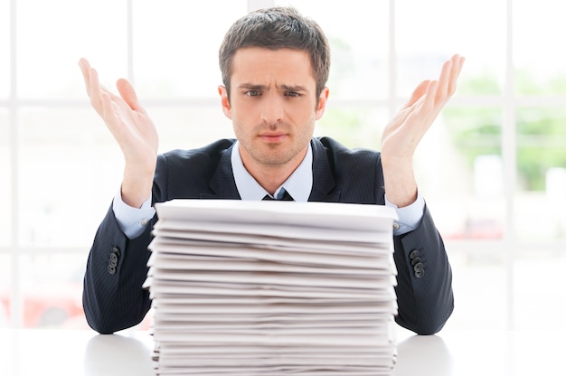 Hopeless businessman. Depressed young man in shirt and tie looking at the stack of paperwork and gesturing while sitting at his working place