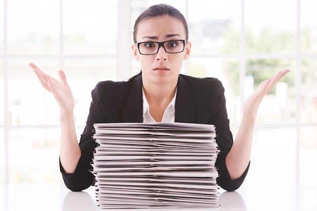 Hopeless businesswoman. Depressed young woman in suit tie looking at the stack of paperwork and gesturing while sitting at her working place