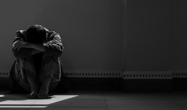 Hopeless man sitting alone with hugging his knees on the floor in empty dark room