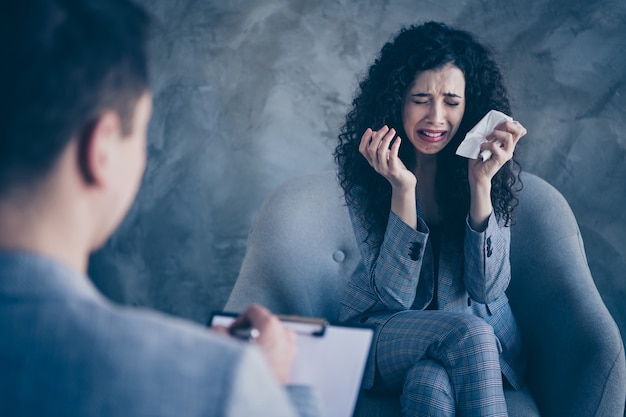 Photo hopeless wavy-haired girl sit in chair talk to psychiatrist cry on grey background