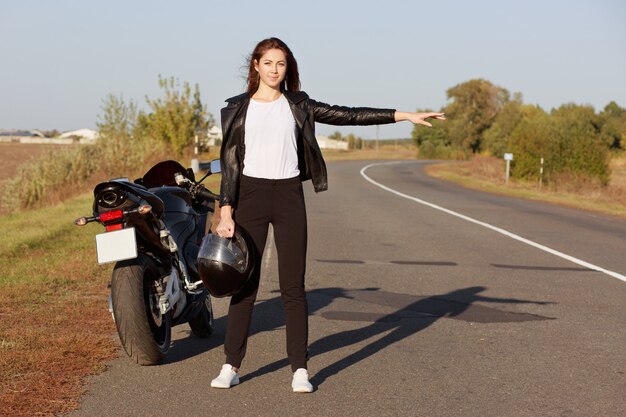 Horiontal-opname van professionele vrouwelijke motorrijder staat in de buurt van brocken-motor, draagt leren jas, houdt helm vast, lift op berm