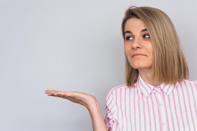 Photo horiontal shot of confused young blonde caucasian female wears pink shirt showing something interesting at copy space on white wall for your information advertising content looking aside in studio