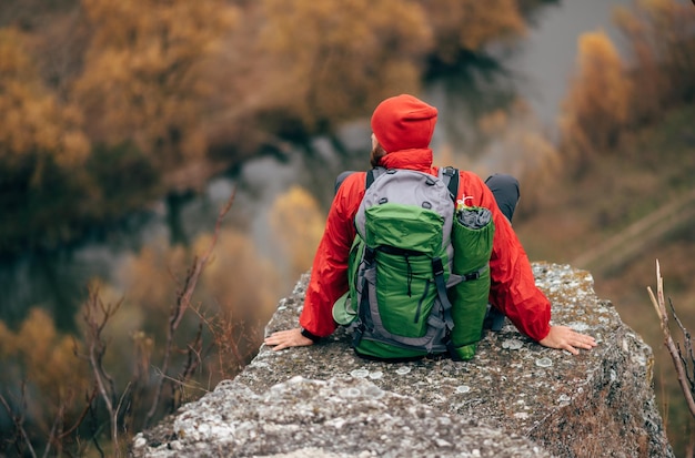 Horizontaal achteraanzicht beeld van jonge knappe wandelaar man zittend op de rots ontspannen en genieten van het prachtige uitzicht op de natuur Reiziger bebaarde man trekking tijdens zijn reis Reizen mensen en levensstijl