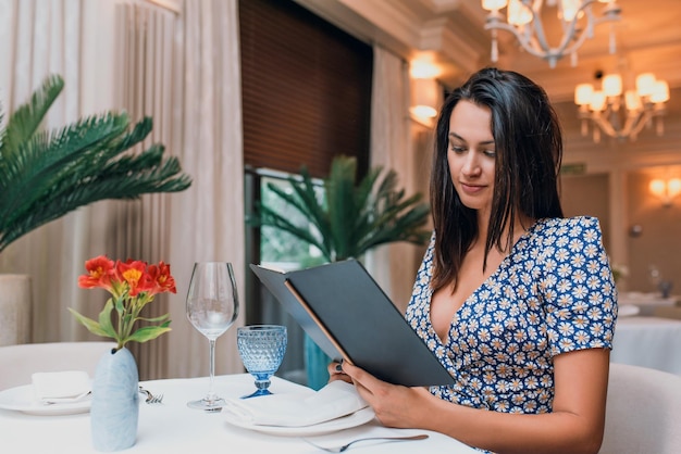Horizontaal beeld van brunette jonge elegante vrouw in jurk zittend in het restaurant met menu voor diner Kaukasische vrouw poseren in cafetaria en wachten op haar vrienden