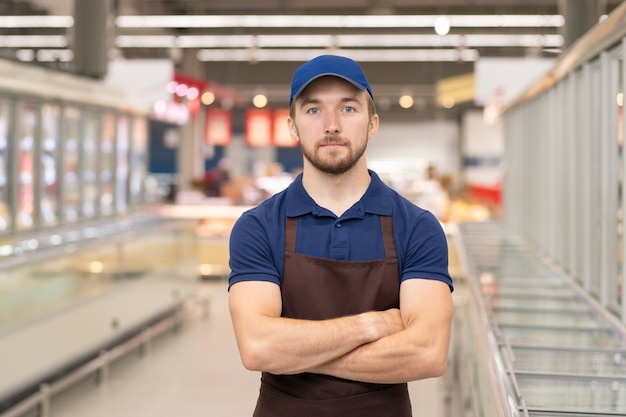 Horizontaal medium portret van een moderne jonge man met een uniform die in de winkel werkt en met gekruiste armen staat