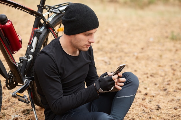 Horizontaal openluchtbeeld van jonge aanhoudende sportmanzitting dichtbij zijn fiets in bos, typend berichten, gebruikend zijn telefoon, aandachtig bekijkend het scherm, dragend zwart uniform voor sport.