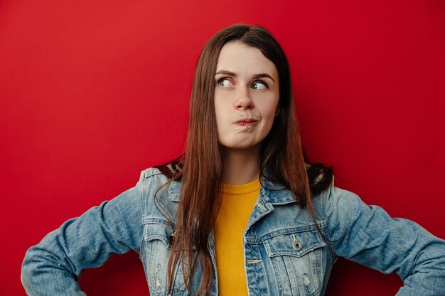 Horizontaal portret van doordachte, verbaasde jonge vrouw die lippen tuit, denkt welke beslissing te nemen, spijkerjasje draagt, besluit in gedachten houdt, staat op rode achtergrond met lege kopie ruimte