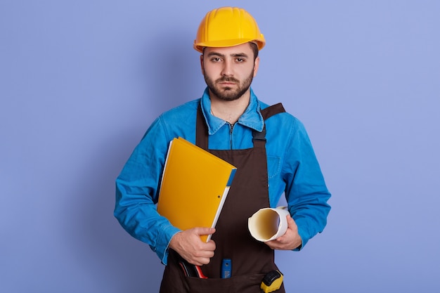 Horizontaal portret van ernstige knappe jonge man met projecten en documenten in beide handen, uniform dragen, ontevreden zijn met de baan. Werkproces concept.