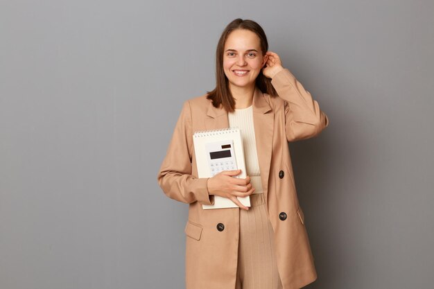 Horizontaal schot van aantrekkelijke vrolijke vrouw, gekleed in een beige jasje in officiële stijl, geïsoleerd over een grijze achtergrond, haar zakelijke documenten en rekenmachine vasthoudend, kijkend naar de camera die haar haar aanraakt