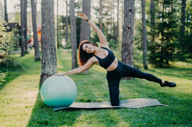 Horizontaal schot van actieve slanke vrouw met vrolijke uitdrukking opzij leunt oefeningen met fitnessbal, gekleed in sportkleding, vormt buiten tegen hoge bomen op karemat. Gezonde levensstijl