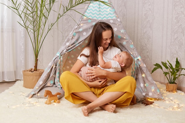 Horizontaal schot van babymeisje in haar moederhanden vrouw spelen thuis met kind in wigwam familie zittend op de vloer op zacht tapijt vrouw die plezier heeft met haar dochtertje