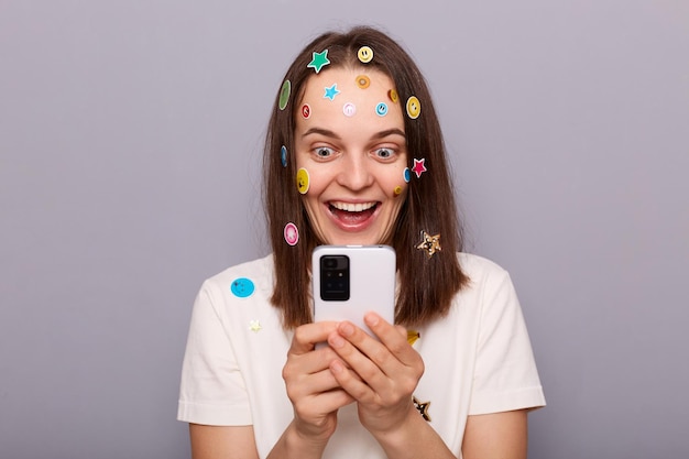 Horizontaal schot van dolblij mooie vrouw bedekt met grappige stickers poseren geïsoleerd over grijze achtergrond sms lezen in mobiele telefoon met goed nieuws kijkend naar telefoonscherm