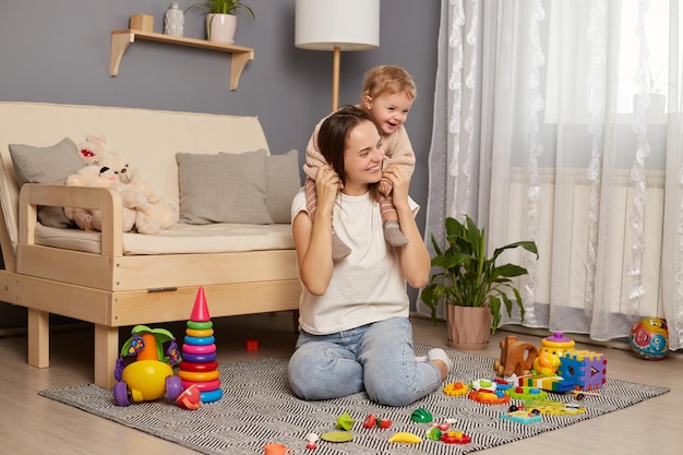 Horizontaal schot van donkerharige vrouw in wit t-shirt en spijkerbroek met haar peuterbaby op haar rug glimlachend zittend op tapijt op de vloer schattig familiespel thuis