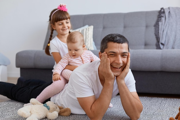 Horizontaal schot van een extreem gelukkige jonge volwassen vader die op de vloer in de buurt van de bank ligt met zijn kleine kinderen op de rug, een man die zijn handen op de wangen houdt en vrolijk lacht, blij om een weekend met kinderen door te brengen.