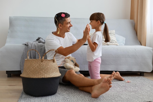 Horizontaal schot van een gelukkige positieve man met een wit t-shirt in casual stijl zittend op de vloer in de buurt van de bank met zijn vrouwelijke kind, familie hand in hand, elkaar met liefde aankijkend.