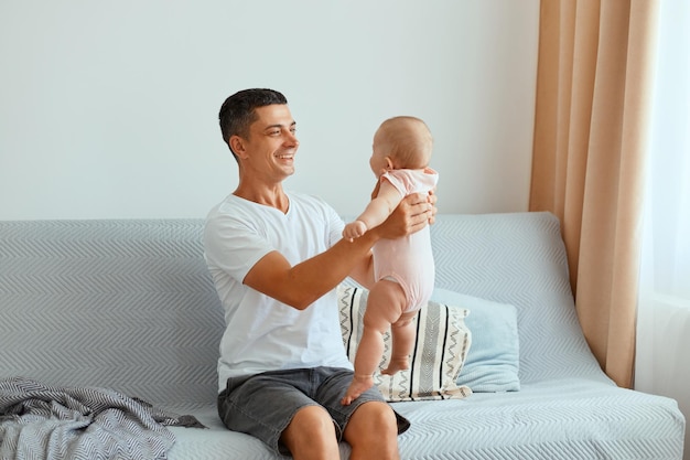 Horizontaal schot van een glimlachende aantrekkelijke man met een wit t-shirt en een spijkerbroek kort zittend op hoest met een schattig klein kind in handen, kijkend naar zijn kind en grote liefde uitdrukkend.