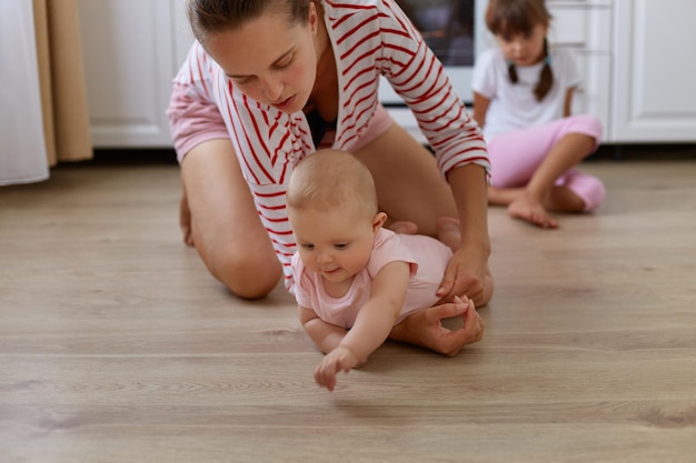 Horizontaal schot van een jonge volwassen vrouw met een gestreept overhemd in casual stijl zittend op de vloer in de keuken met kinderen ouder kind zittend in de buurt van oven moeder spelen met kruipende baby baby