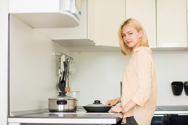 Horizontaal schot van een mooie blonde studente die in de buurt van een fornuis staat en voedsel kookt in een koekenpan in een witte keuken, kijkend naar de camera. Thuis koken.