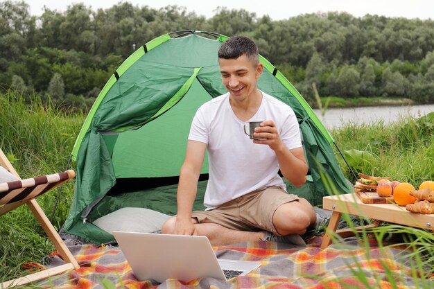 Horizontaal schot van een opgetogen blanke freelancer die in de buurt van de rivier in zijn tent zit en online aan een notebook werkt terwijl hij koffie of thee drinkt en met een glimlach naar een laptopscherm kijkt