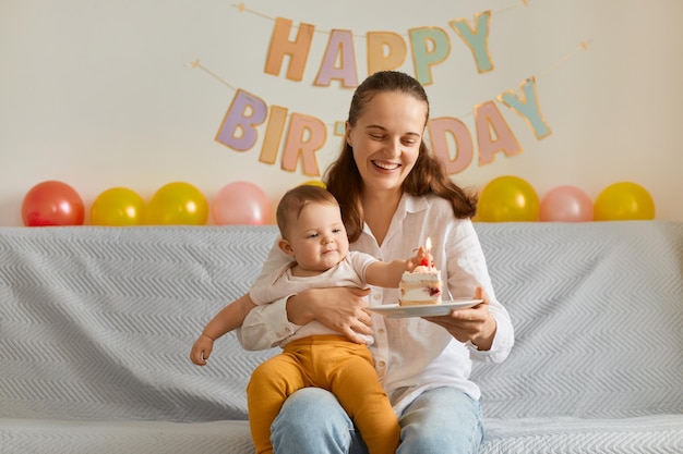 Horizontaal schot van een positieve glimlachende vrouw die met haar peuterdochter op de bank zit en cake vasthoudt met kaarsen die geluk uitdrukken bij het vieren van de eerste babyverjaardag