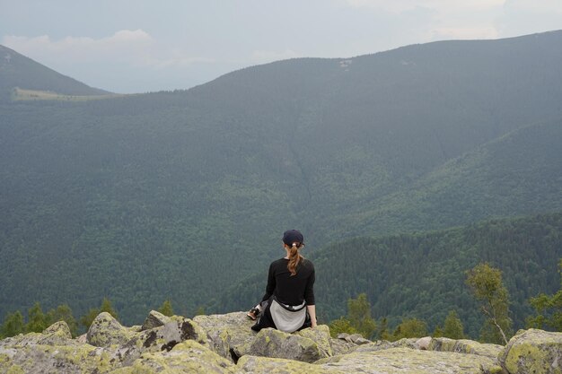 Horizontaal schot van een vrouw die op een open haard zit en na het wandelen rust en kijkt naar een prachtig uitzicht op de bergen en het achteraanzicht van het bos