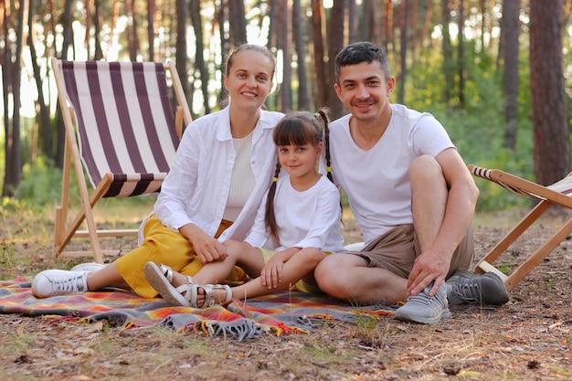 Horizontaal schot van familie met witte overhemden die op een deken in het park zitten en met een glimlach naar de camera kijken blanke ouders met kleine dochter die samen in het bos ontspannen