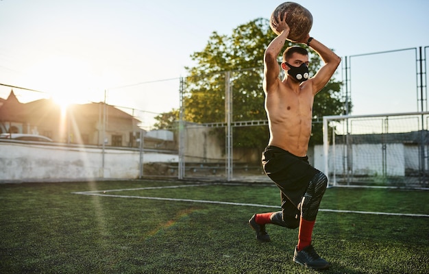 Horizontaal schot van fit gezond gespierd mannetje dat oefeningen doet met medicijnbal in masker in stadion buitenshuis Kopieer ruimte voor reclametekst Shirtless sportman doet training buiten op sportveld