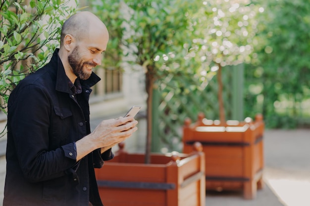 Horizontaal schot van gelukkige kale mannelijke hipster met baard gekleed in zwarte jas houdt slimme telefoon leest informatie of nieuws op webpagina geniet van gratis internetwandelingen buiten in prachtig park