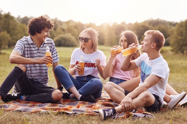 Horizontaal schot van gelukkige vrienden hebben picknick, drinken koud bier, genieten van zomerweer, zitten op plaid, delen laatste nieuws, poseren tegen groene natuur