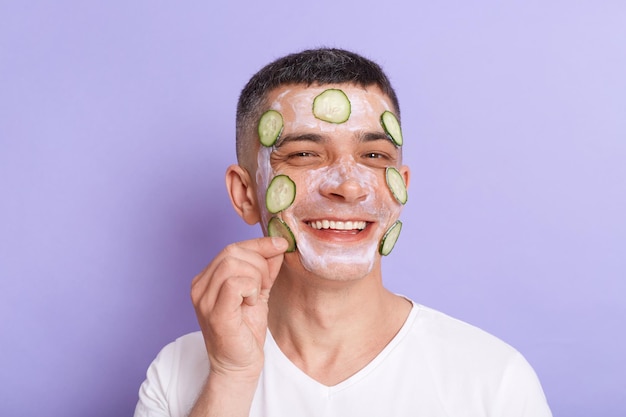 Horizontaal schot van glimlachende tevreden man met wit t-shirt die masker met plakjes komkommer op zijn gezicht toepast, genietend van cosmetologie huidverzorgingsprocedures geïsoleerd over paarse achtergrond